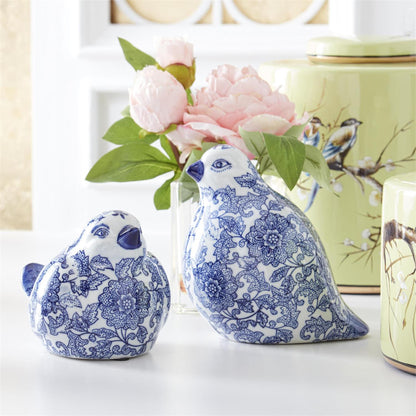 2 styles of blue and white patterned birds on a table with a vase of pink flowers behind them.
