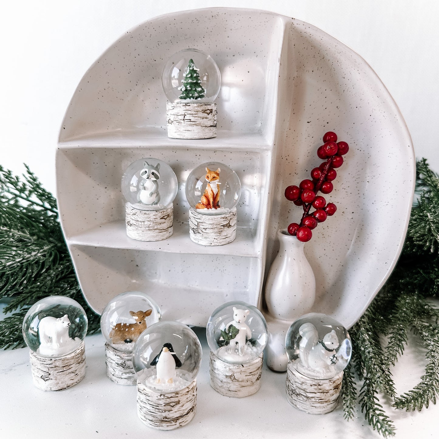 White, flecked ceramic shelf featuring water globes with animals inside. Faux greenery in the background and red berries in a tiny vase.