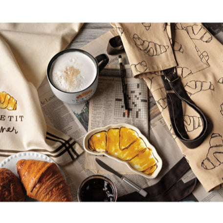 Breakfast setting with coffee, croissant shaped dish, and fruit on a newspaper