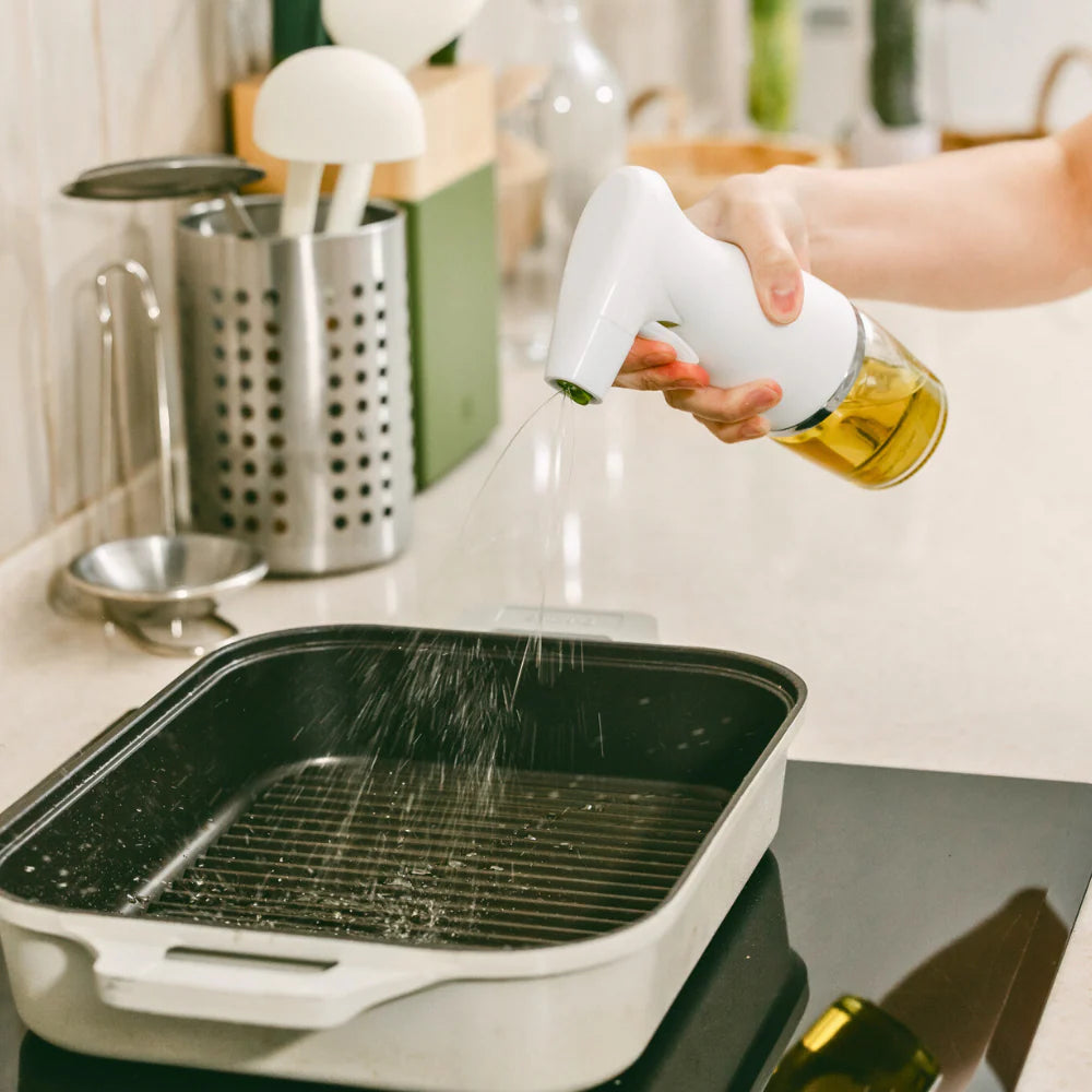 Person spraying oil into a black grill pan on a kitchen counter.