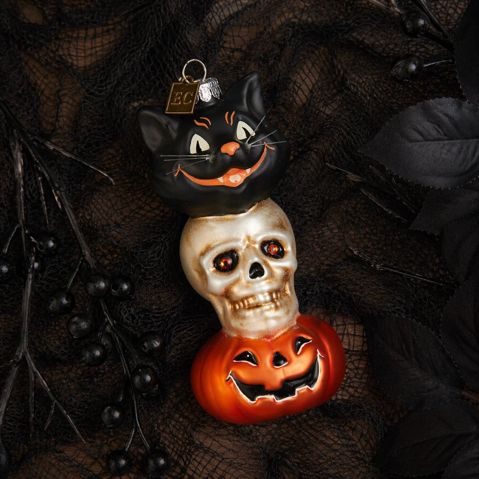 A stacked Halloween glass ornament featuring a black cat on top of a skull, which is on top of a carved orange pumpkin, styled on a dark black lace fabric background.