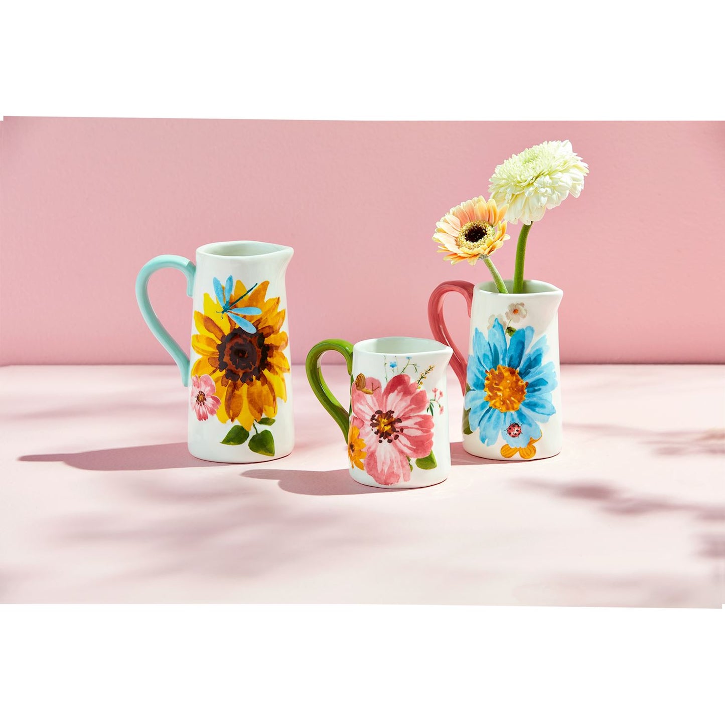 3 styles of floral and bug pitcher vases arranged on a pink background, the medium vase has daisies in it.