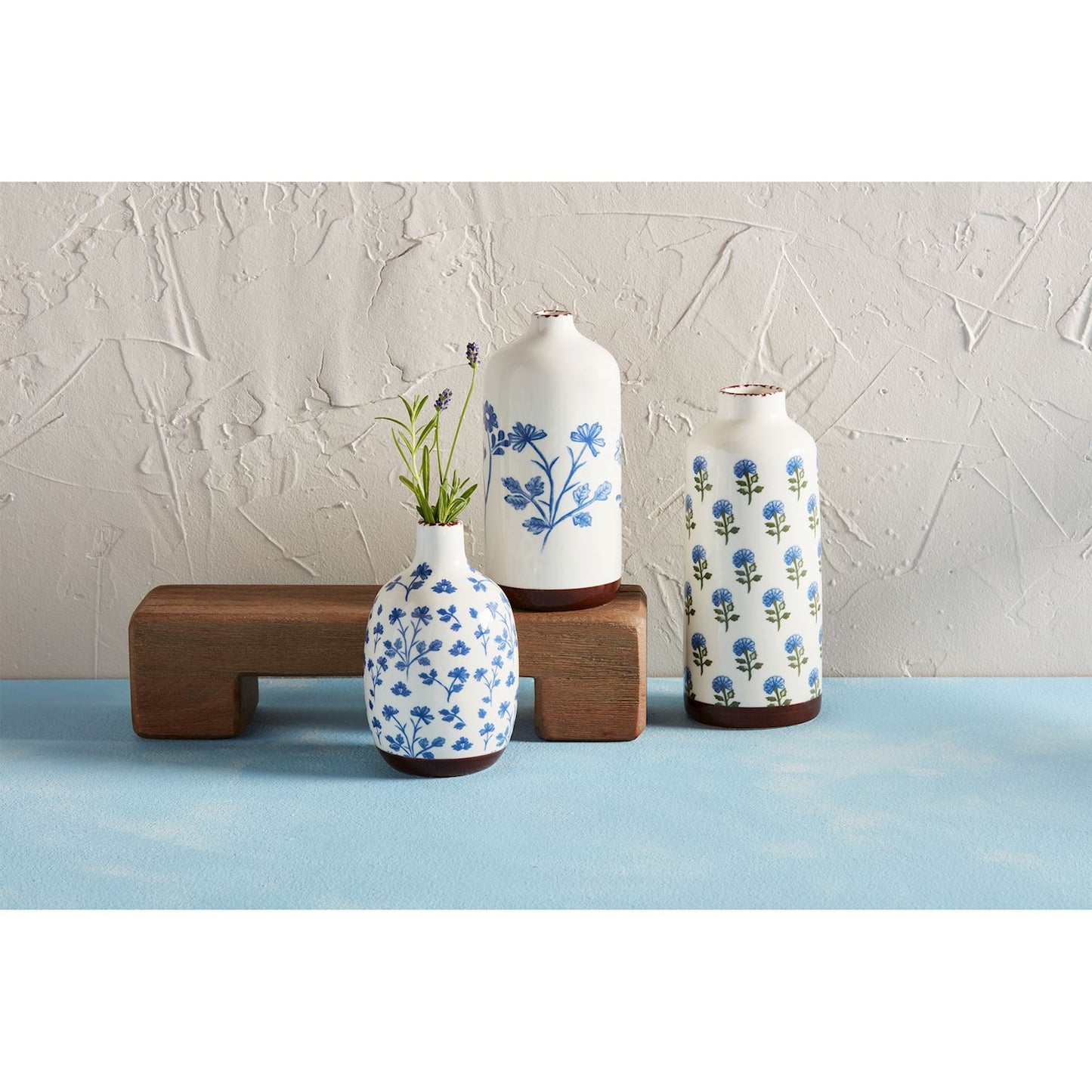 3 sizes and style of blue floral vases arranged on a blue table with a riser, small vase has greenery in it.
