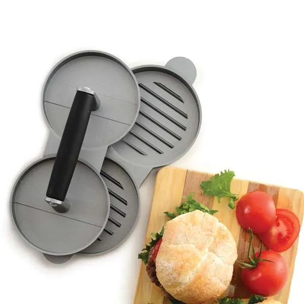 Top view of a white surface with the double burger press and a wooden board with a burger and sliced tomatoes on it