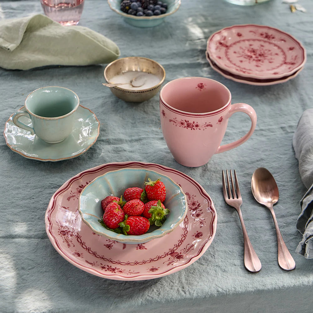 Table setting with pink ceramic dishes, including a mug and bowl of strawberries, on a table cloth