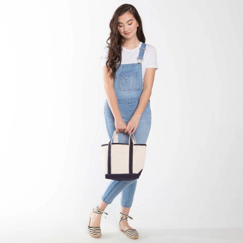 person wearing denim overalls and a white shirt holding off white tote with navy handles and trim