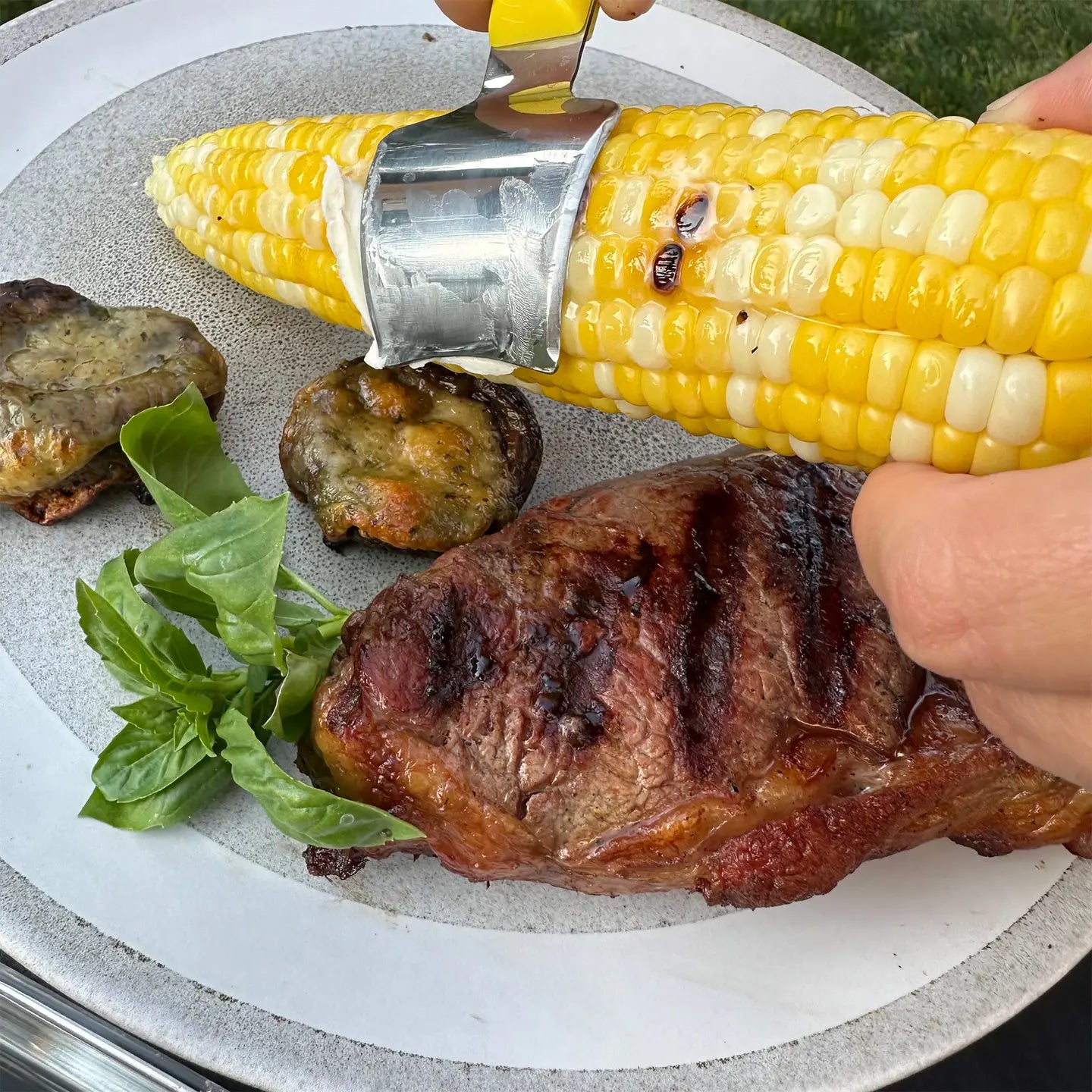 Grilled corn on the cob bring buttered with corn butter knife held over a plate with grilled meat and vegetables.