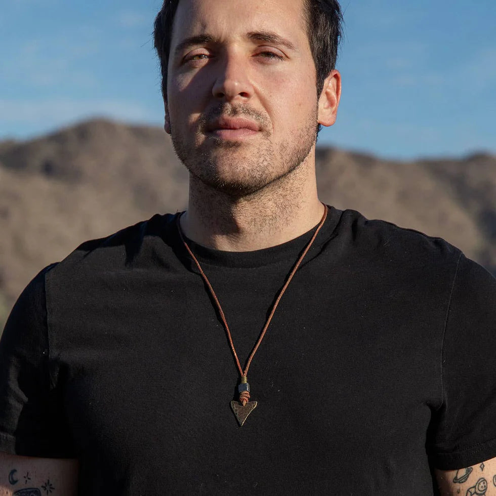 Man wearing a black shirt with a necklace, standing against a mountainous landscape.