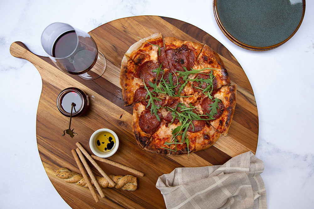 top view of altea serve board with a pizza, bowls of oil, bread sticks, and a glass of wine on it.