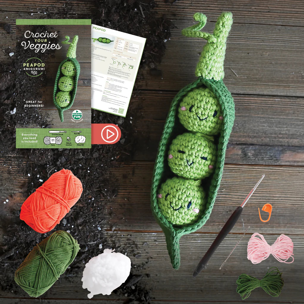 Crocheted green peas in a pod with yarn, needles, and packaging on a wooden surface.