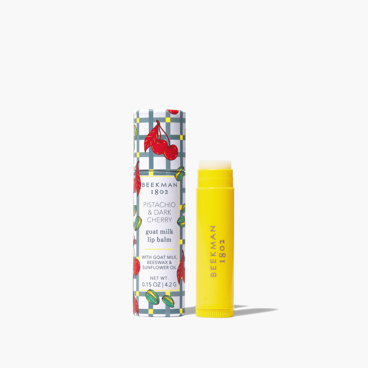 yellow tube of Pistachio Dark Cherry Lip Balm set next to its tube packaging with a cherry and check design