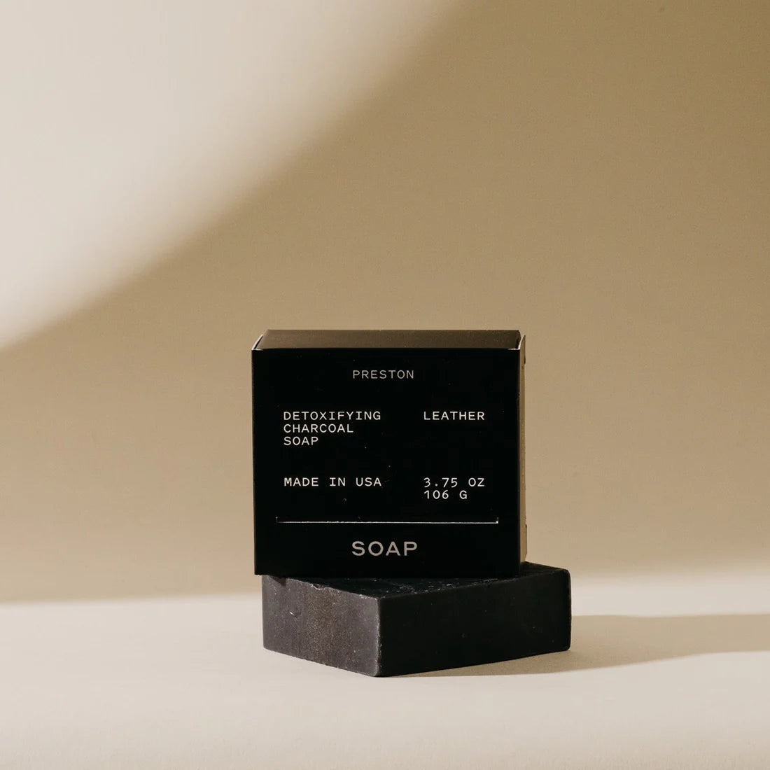 Black bar of soap with packaging set on top of it on a beige background