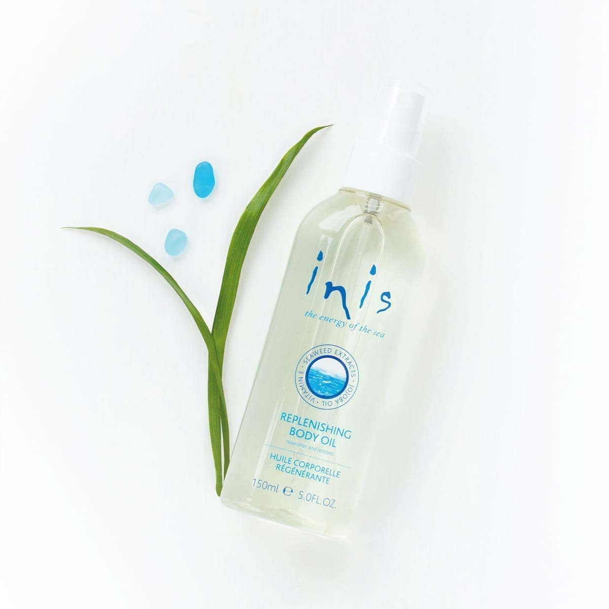 spriy bottle of inis body oil arrangedw ith seaweed and blue sea glass on a white background.