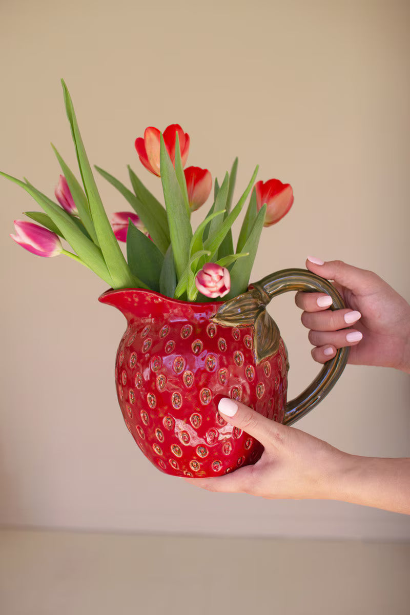 Red pitcher filled with red and pink tulips held by a hand against a beige background