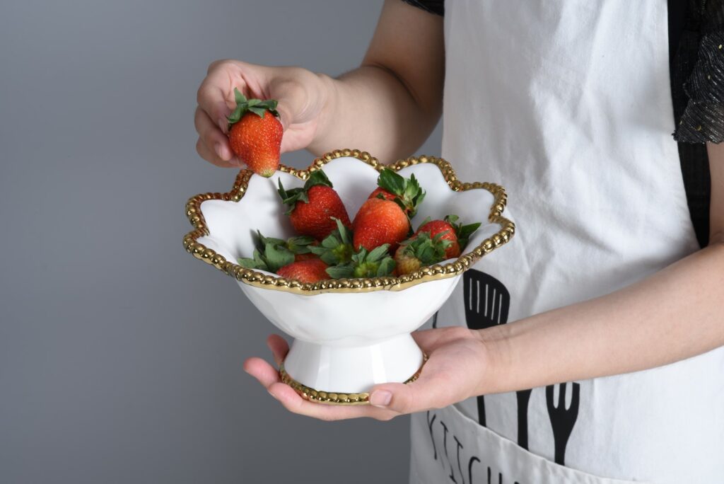 hands holding white footed bowl with gold beaded rim filled with strawberries