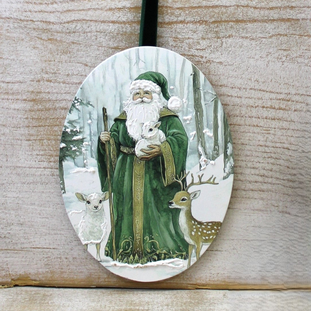 An oval Christmas ornament featuring a painted illustration of Saint Nicholas in a green robe holding a white rabbit, with a deer and lamb beside him in a snowy forest. The ornament has a dark green hanging loop and is shown against a light wood background.