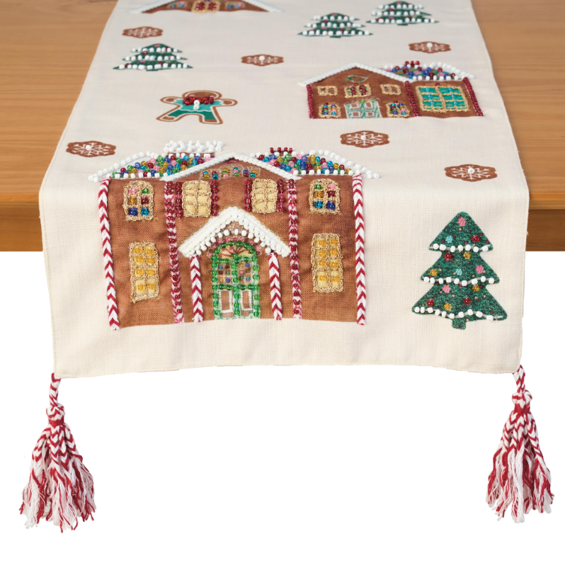 A cream-colored table runner with embroidered gingerbread houses, Christmas trees, and candy canes, draped over a wooden table with red and white tassels hanging from the ends.