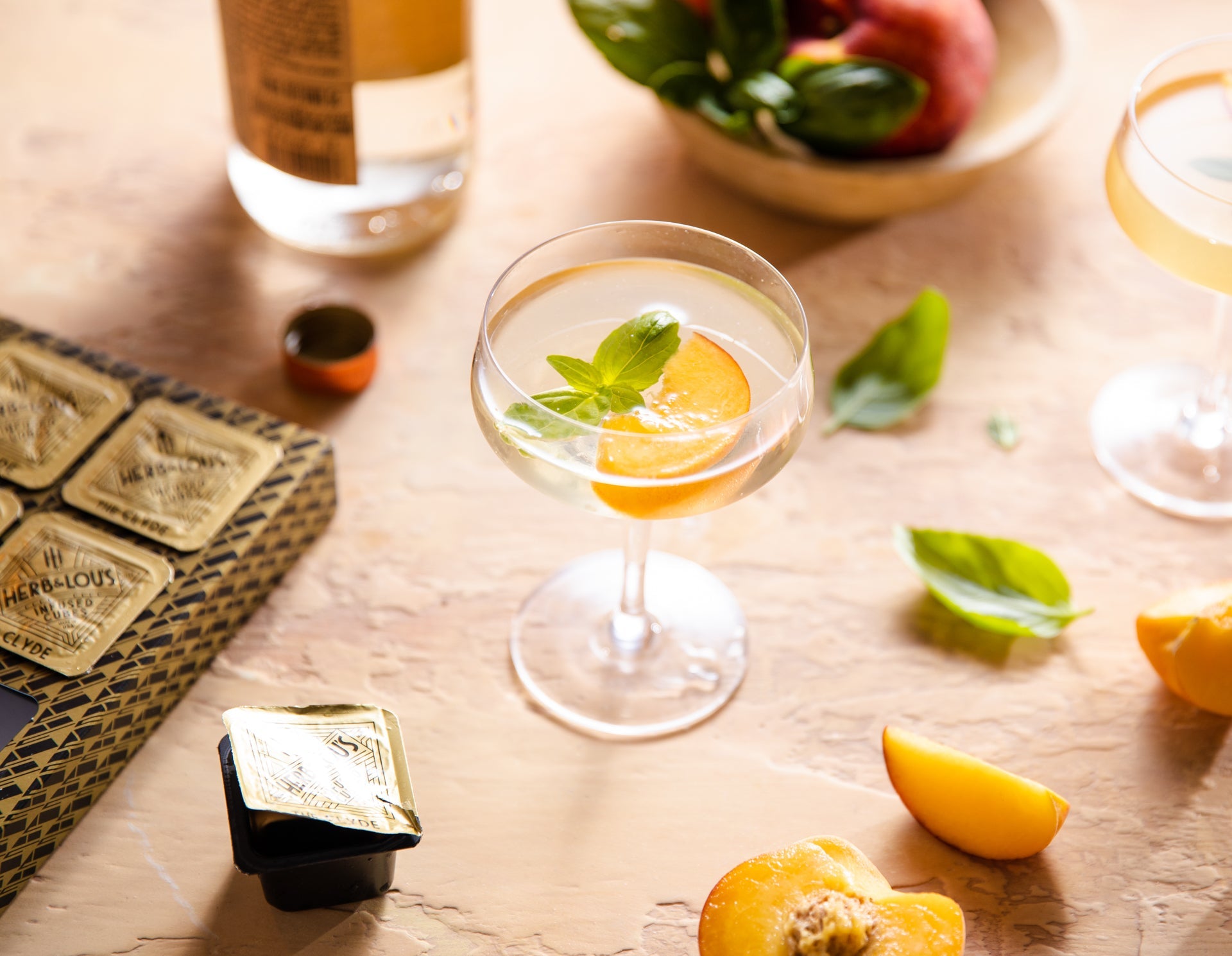 glass filled with Peach Cosmopolitan with Benedictine-Inspired Herbs & Artisanal Bitters arranged with peaches, leaves, bottles, and glasses.