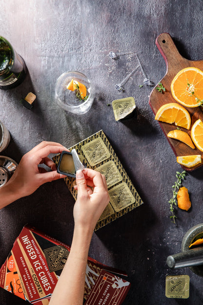 The Cooper 6 Cube Pack on a slate background with hand opening a frozen cube arranged with orange slices, herbs, bottles, and glasses.
