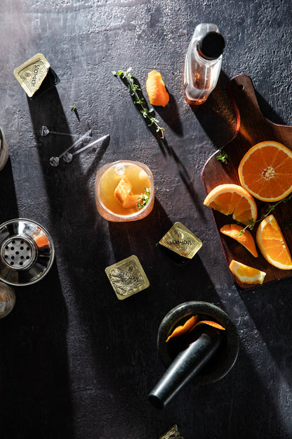 The Cooper cubes on a slate background arranged with orange slices, herbs, bottles, and glasses.