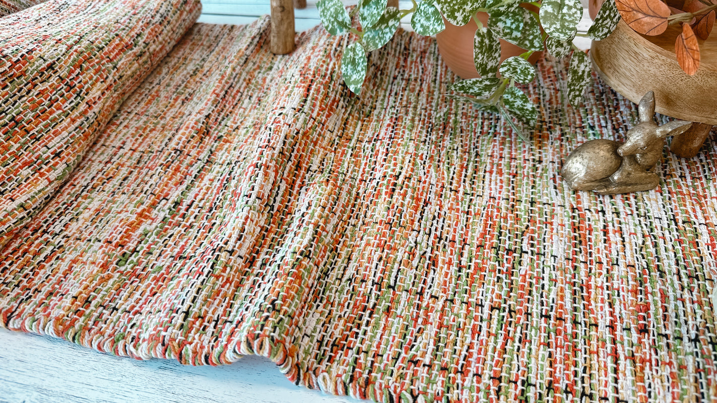 A close-up of a woven textile runner with a multicolor blend of orange, red, green, beige, and grey tarns, styled with a potted plant and a small gold deer figurine.