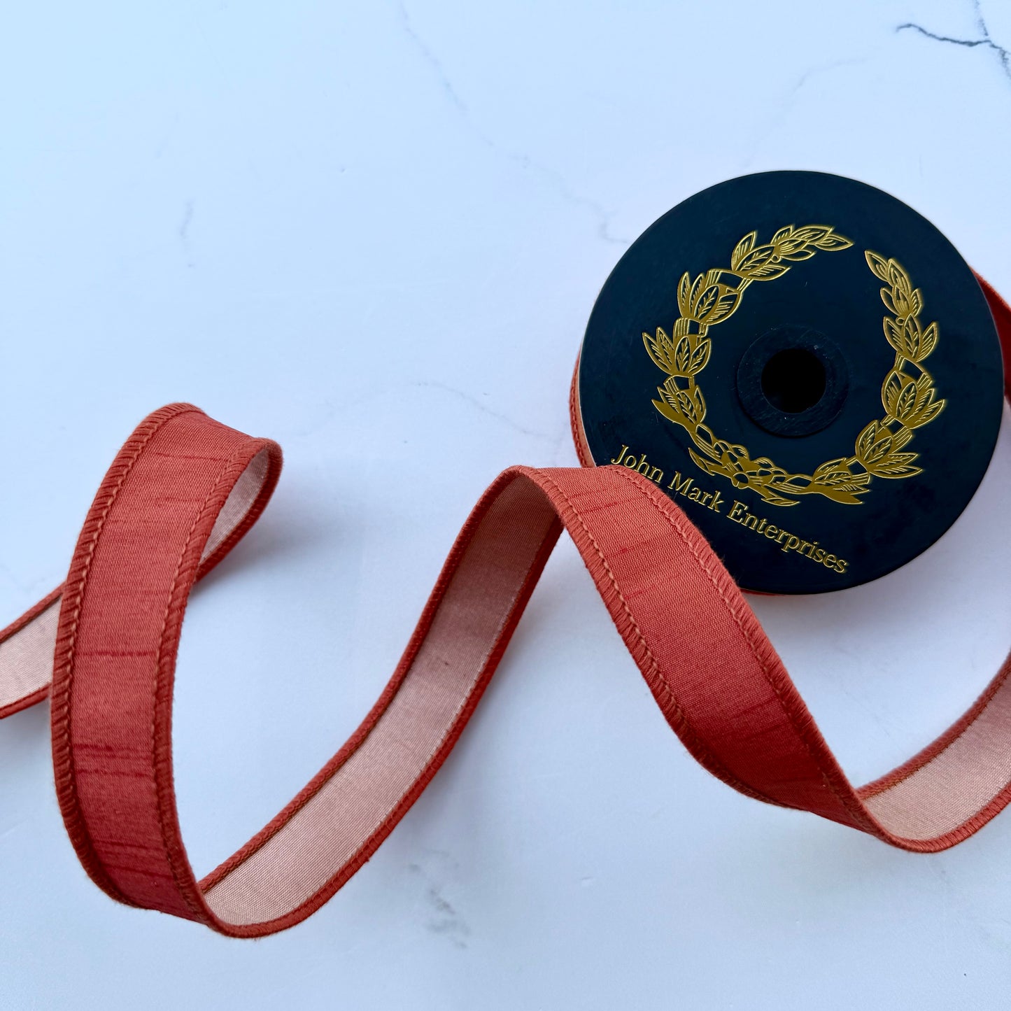 orange ribbon with light orange backing unspooled from a black spool with gold foil branding, set against a light background.