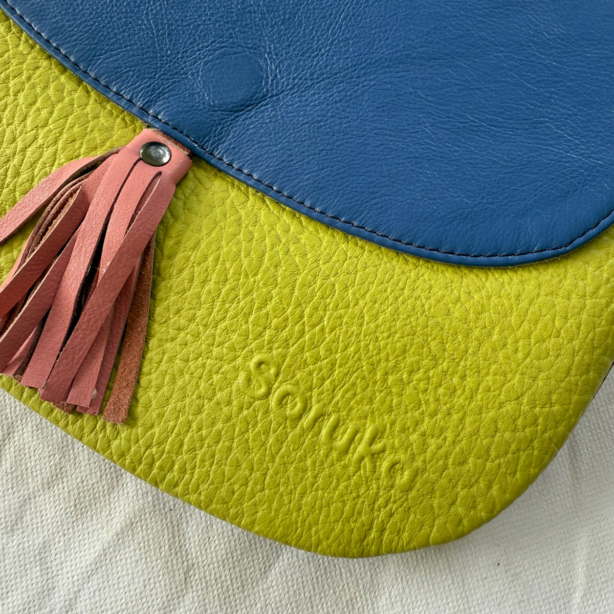 A close-up of a textured leather shoulder bag with a bright green body, a blue flap, and a dusty pink leather tassel with a silver metal rivet