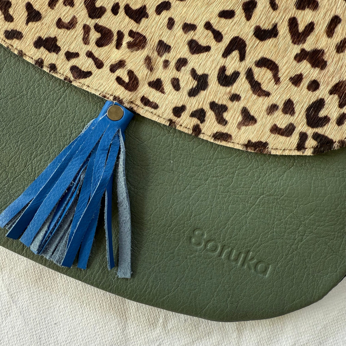 Close-up of a Soruka bag showing a blue leather tassel, a beige and brown leopard print flap, and an olive green textured body with the brand name embossed.