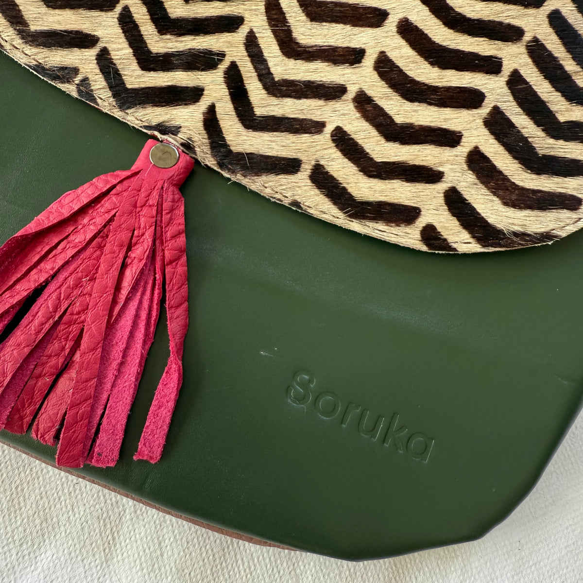 Close-up of a Soruka bag featuring a dark green leather base, a pink leather tassel, and a beige and black chevron-patterned leather flap.
