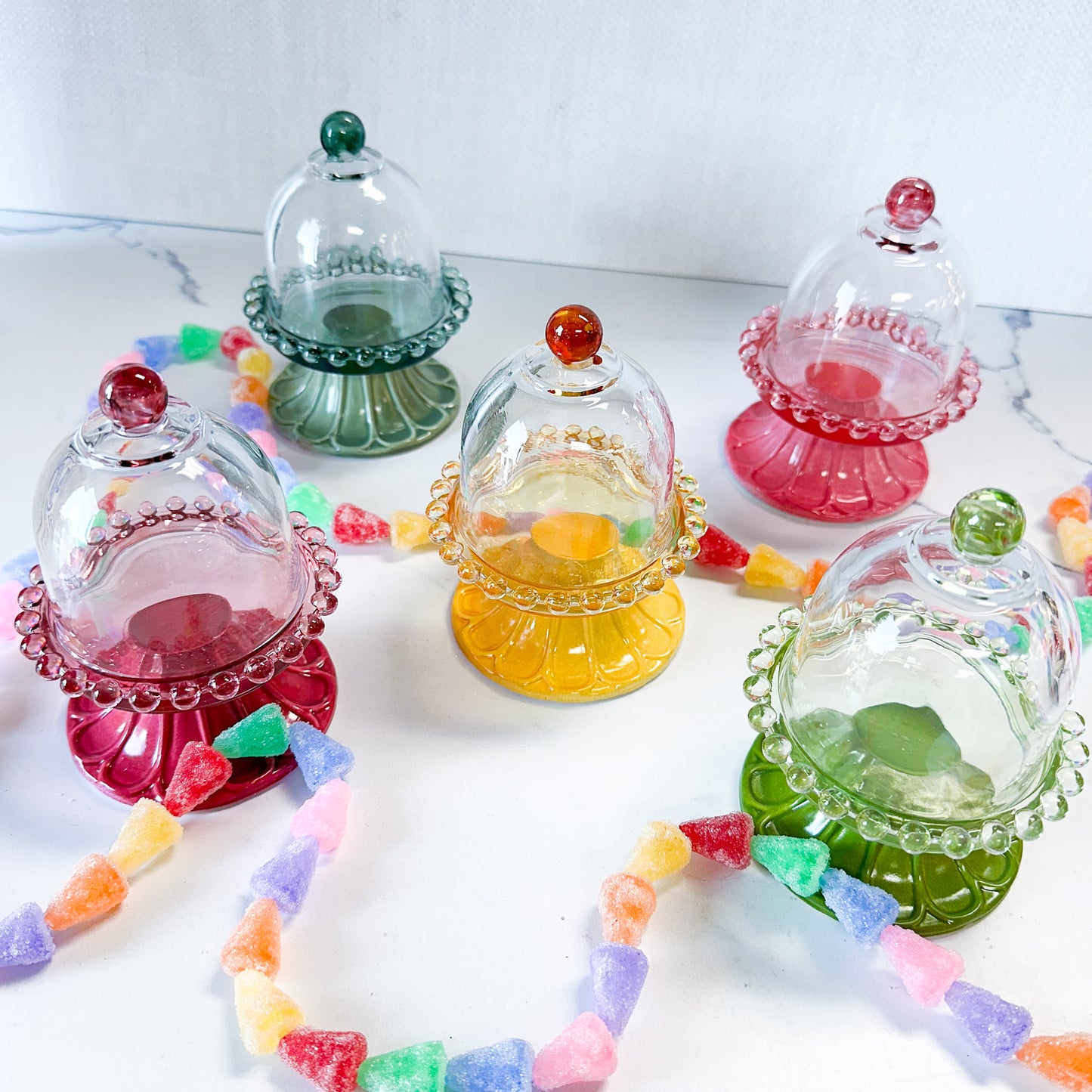 Colorful glass pedestals with cloches arranged on a white surface with a strand of gun drop candies