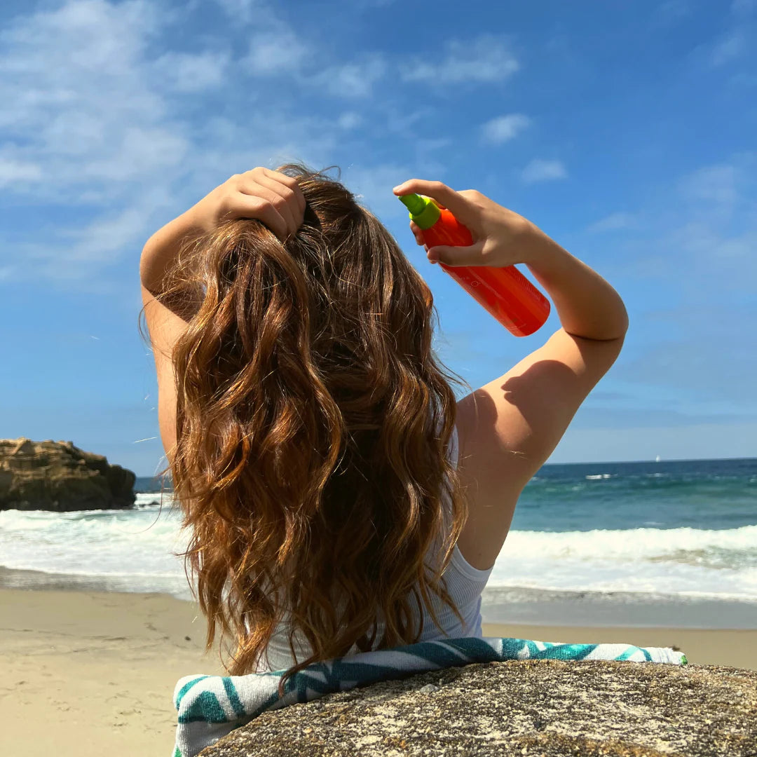 Person applying leave-in conditioner on a beach with ocean and sky in the background