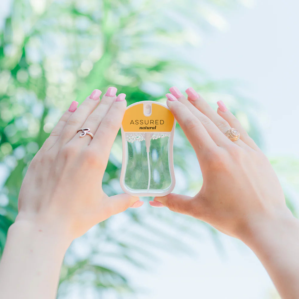 Hands holding a 'Assured' body mist bottle against a blurred green background