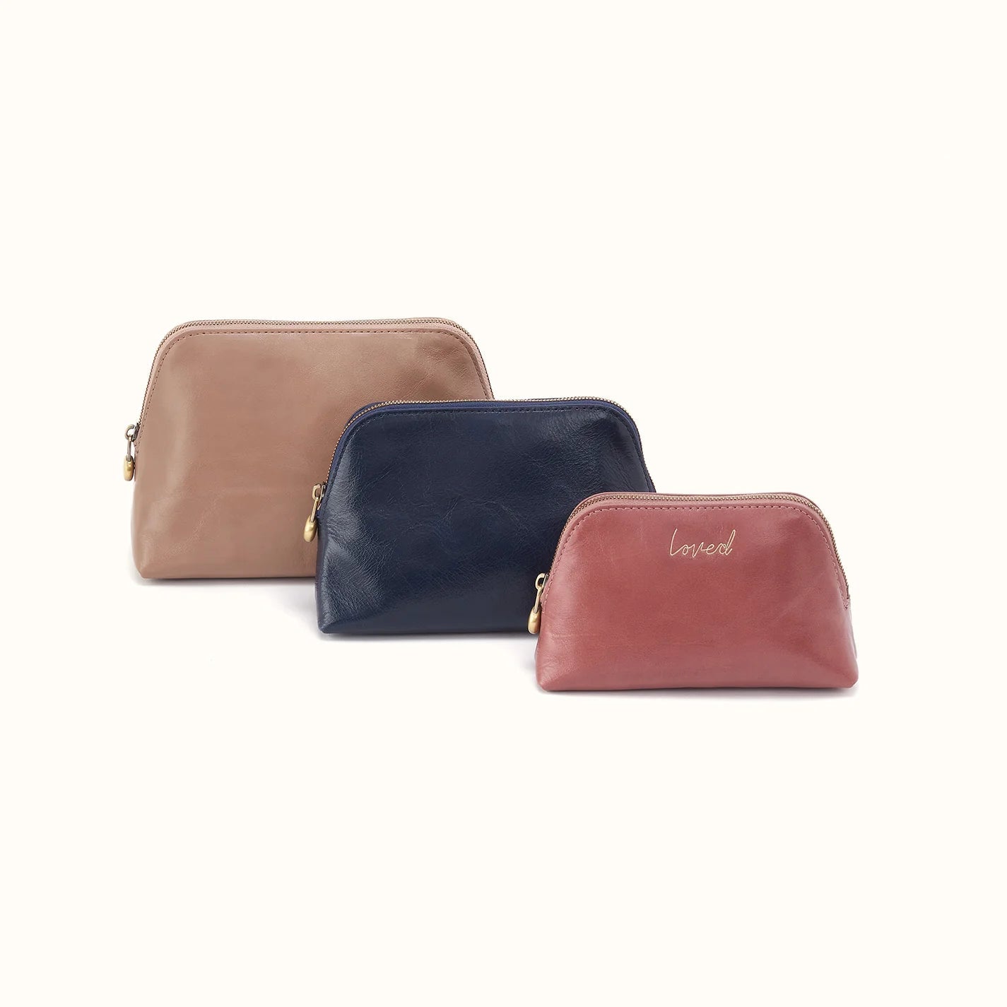 tan pouch behind a navy pouch behind a rose color pouch that has gold script on it