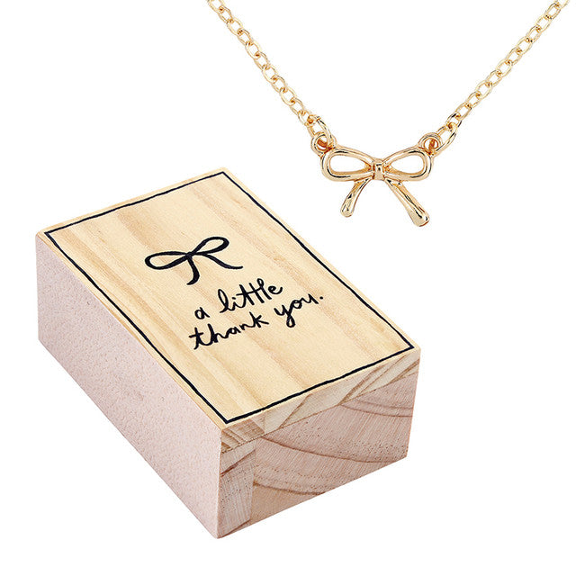 Gold necklace with bow detail next to a wooden box with 'a little thank you' text.