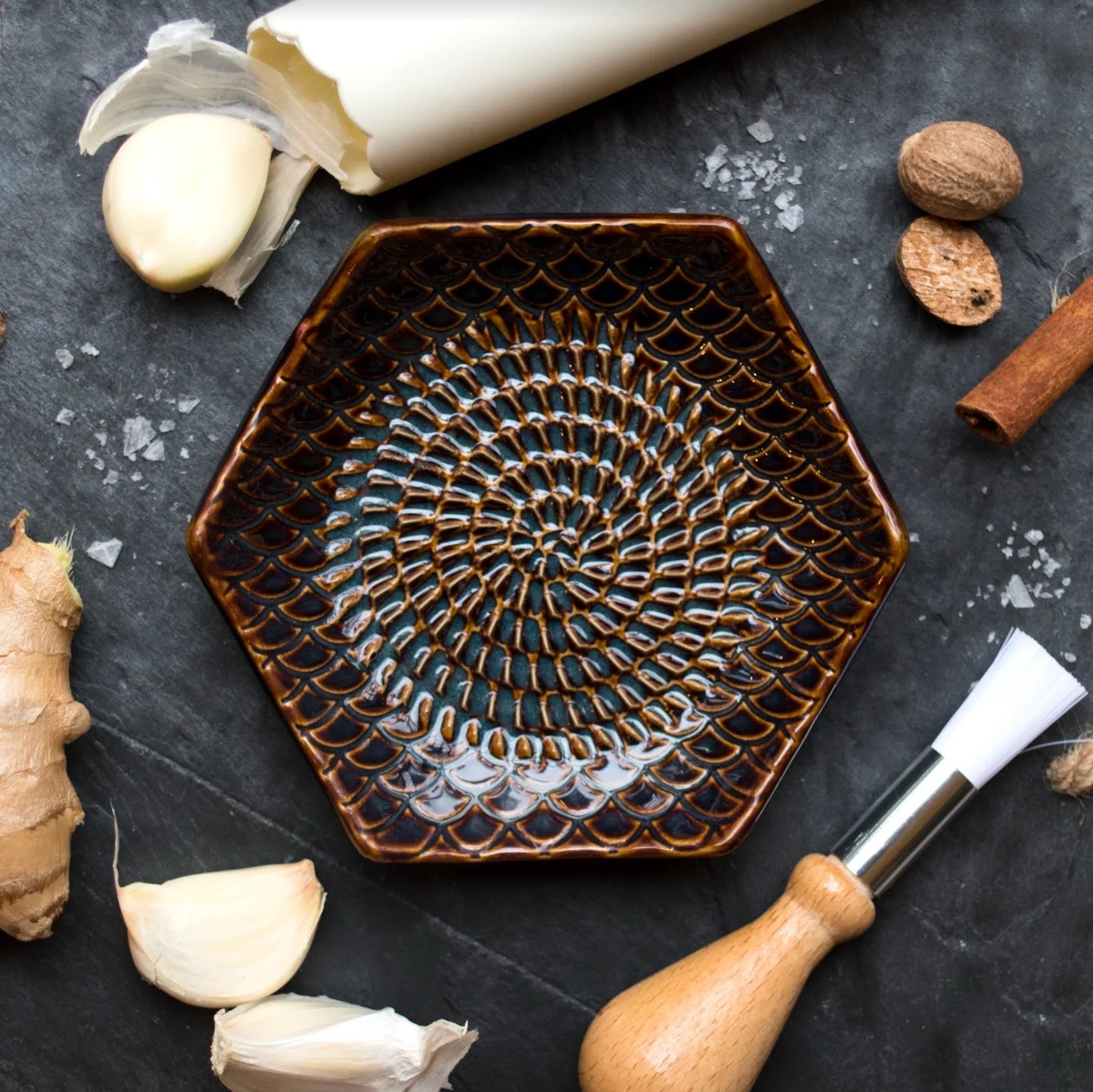 top view of a black countertop arranged with the tortoise grate plate, silicone garlic peeler, brush, and a variety of spices and roots.