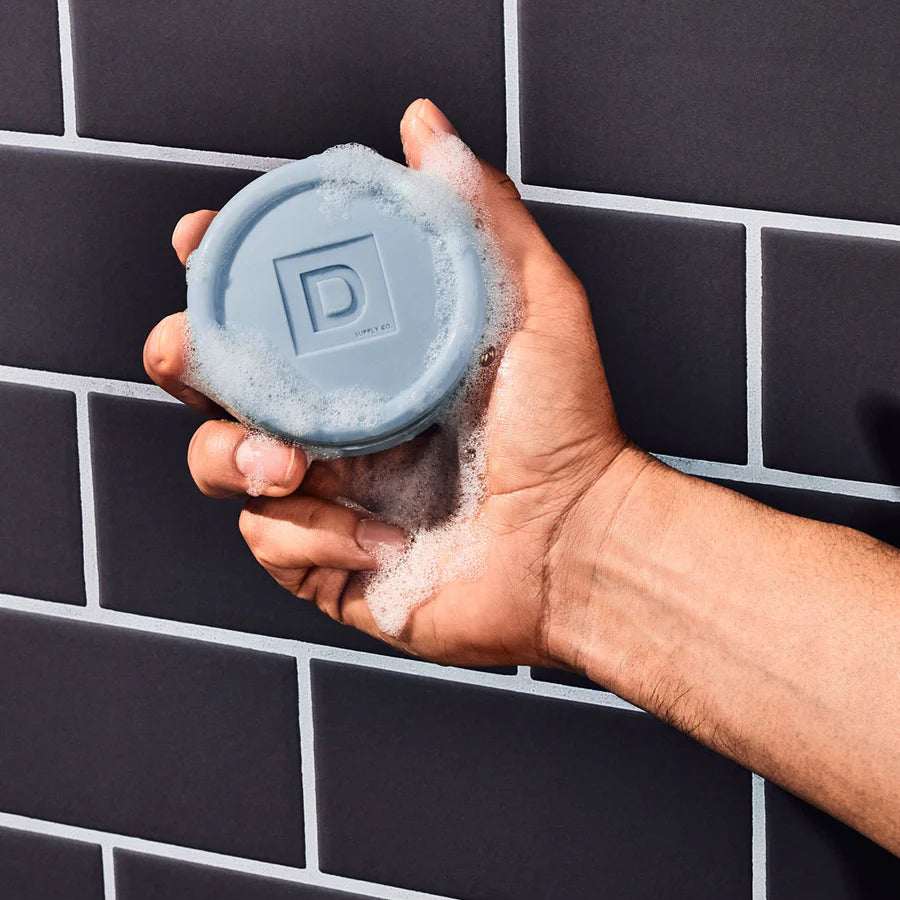 soapy Hand holding a blue bar of soap with a brand logo against a black tiled wall background