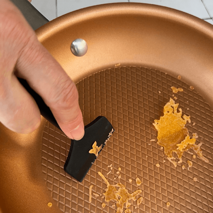 hand using a black tool to scrape food residue from a copper-colored frying pan.