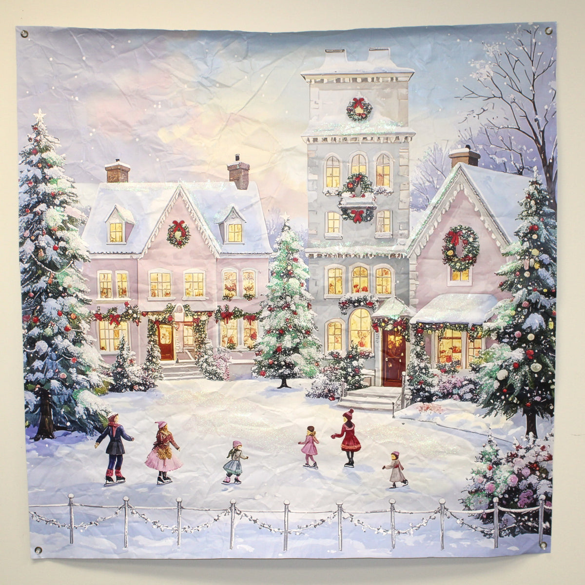 A square wall art print showing a snowy winter village scene with large decorated houses, Christmas trees, wreaths, and children ice skating in the foreground. The artwork fills most of the frame and is photographed against a plain light background.
