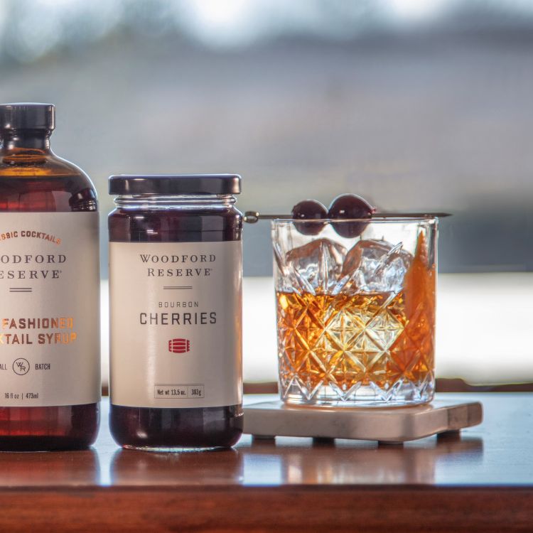 bottle of  Woodford Bourbon Cherries on a table with a cocktail with 2 cherries on a skewer in the glass.