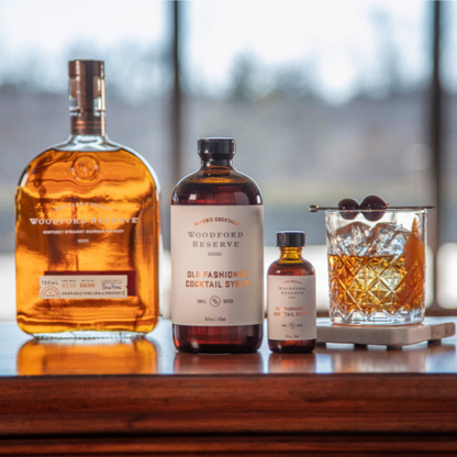 2 sizes of Woodford Reserve Old Fashioned Cocktail Syrup on a table with a bottle of bourbon and a cocktail.