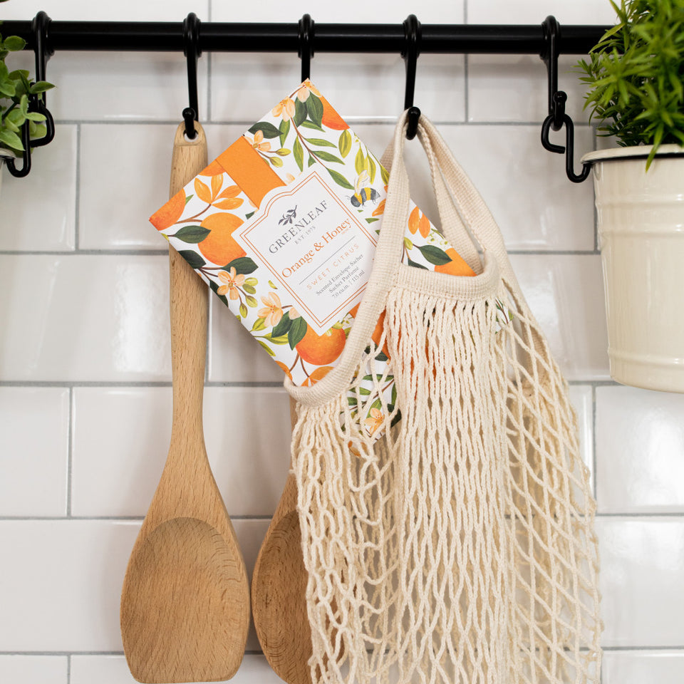 Orange & Honey Large Sachet tucked into a mesh bag hanging with utensil against a subway tile backsplash.