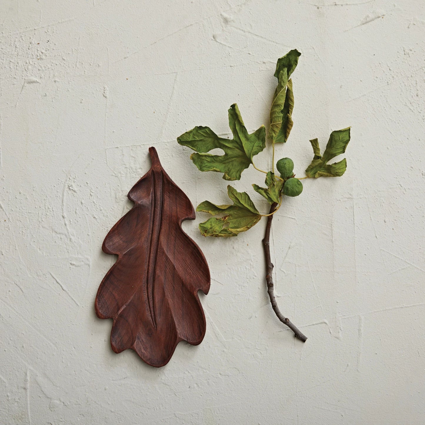 top view of wood leaf shaped board set next to a fig branch on a plaster surface.