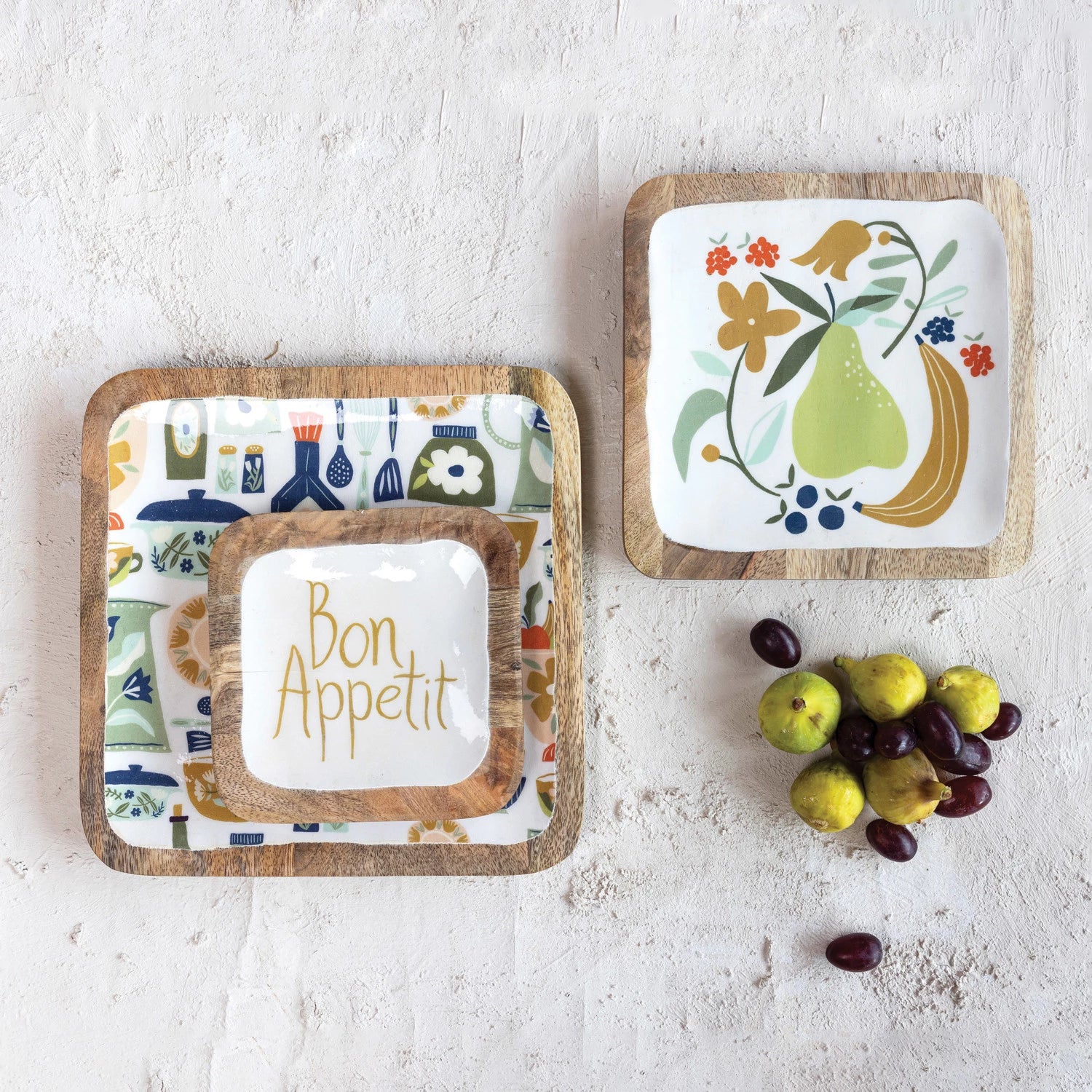 3 styles of enameled mango wood trays arranged on a plaster surface with figs and olives.