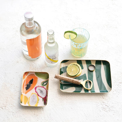 2 styles of Enameled Metal Tray with Abstract Design arranged on a counter with bottles and a glass, the large tray has lime wedges and a bottle opener on it.