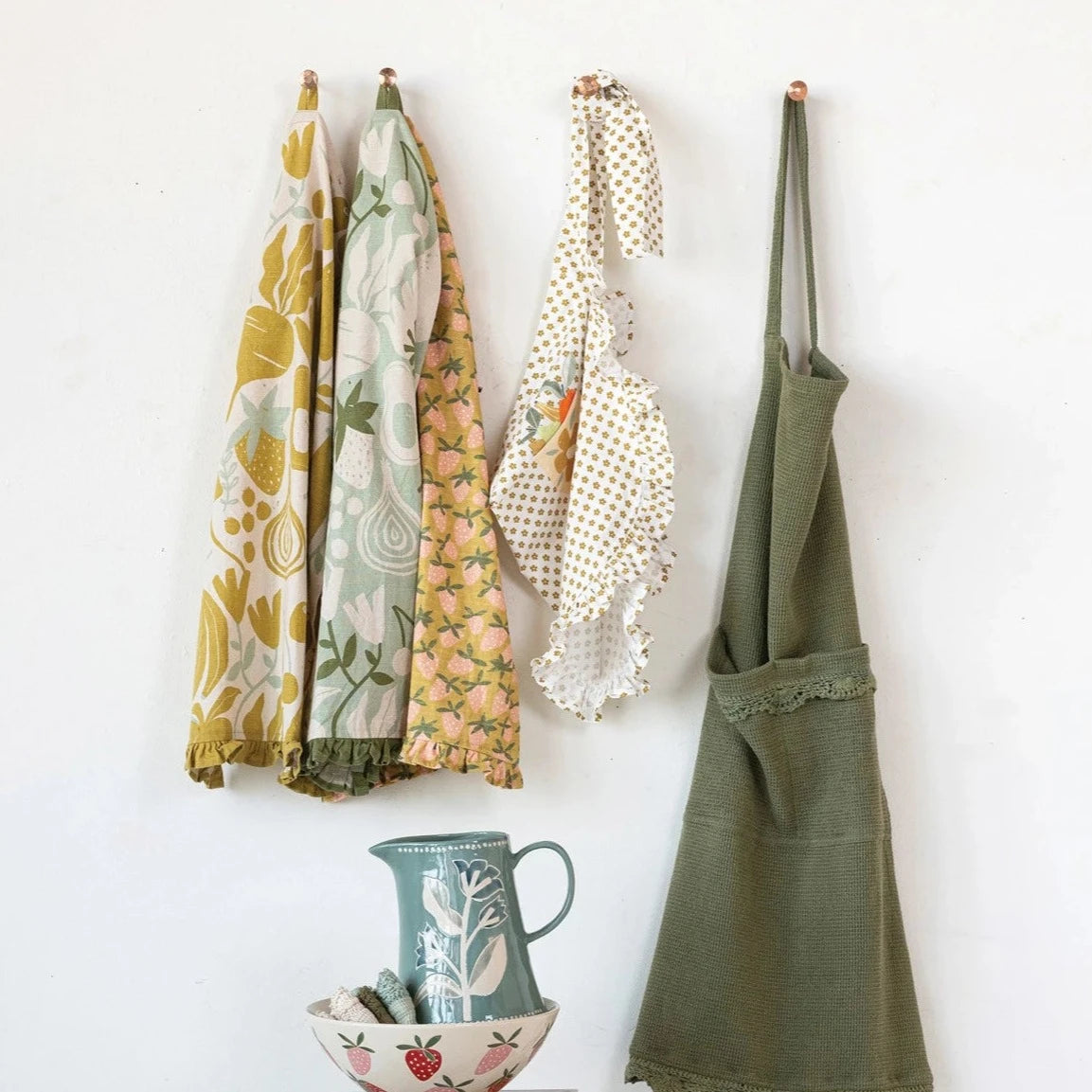 3 styles of fruit and veggie tea towels hanging on hooks next to aprons hanging on other hooks and a bowl and a pitcher stacked underneath.