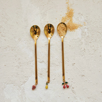 3 styles of spoons with either a strawberry, pineapple, or cheery charm on each end arranged on a plaster background with brown sugar in one spoon.