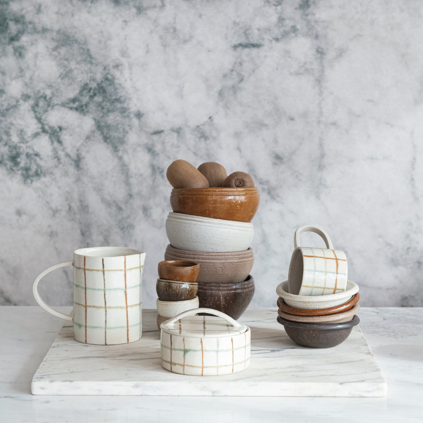 stacks of dishes and mugs arranged with a plaid pitcher on a marble countertop.