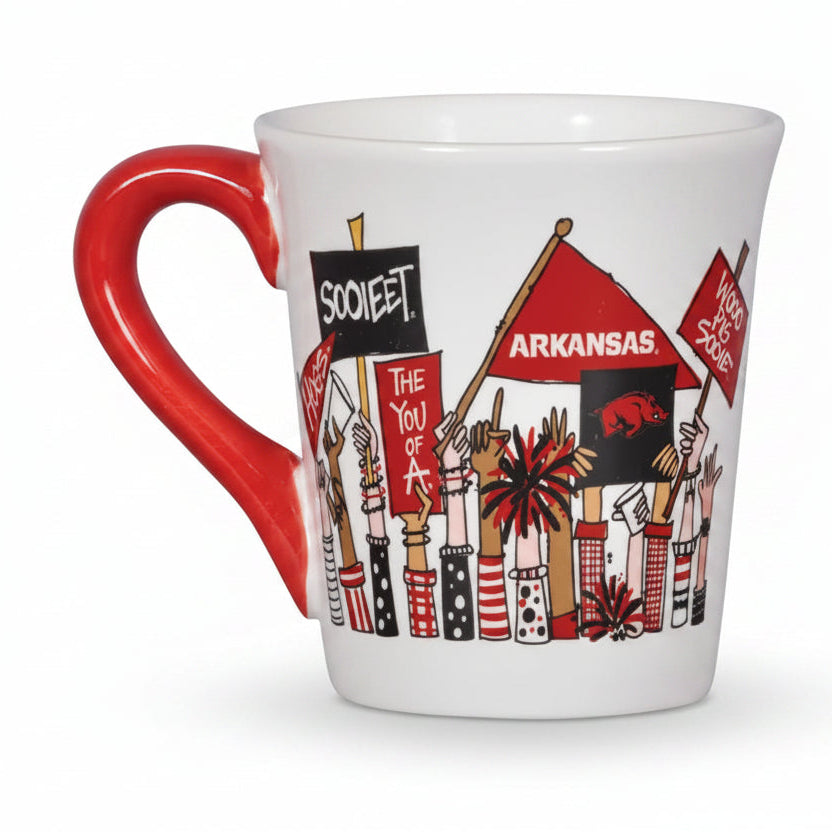 A white ceramic mug with a bright red handle, decorated with an illustration of fans holding Arkansas flags and signs, including 'sooie' and 'ARKANSAS'.