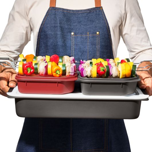 a man wearing a denim apron holding the good grips grilling prep and carry system with cabobs on top