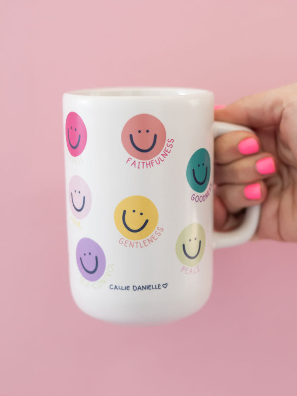 White mug with smiley face designs and text held by a hand on a pink background